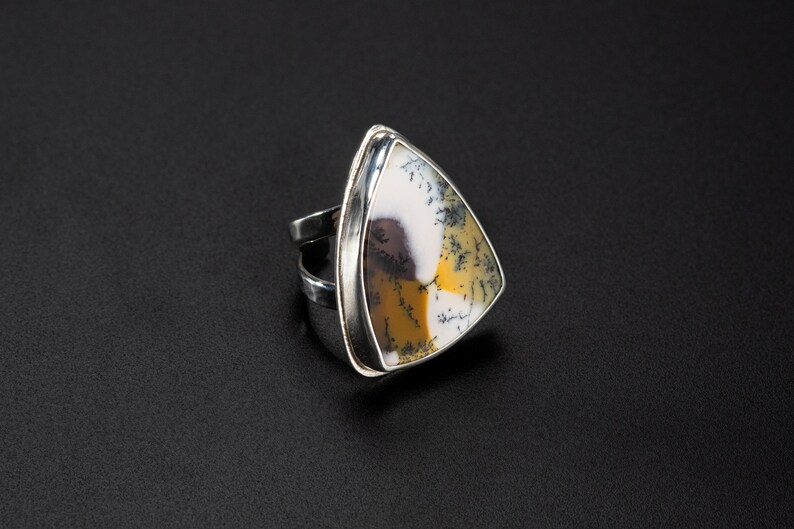 Puede incluir: Un anillo de plata con una piedra triangular grande. La piedra presenta un patr&oacute;n &uacute;nico de blanco, amarillo y negro, engastada en una banda de plata pulida. El anillo es una pieza llamativa.