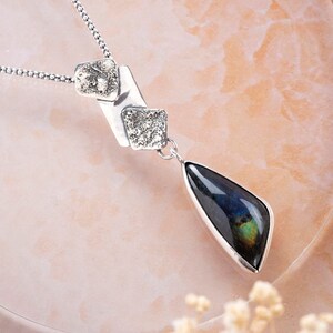 May include: A silver necklace with a teardrop-shaped pendant featuring a dark blue labradorite gemstone. The pendant is attached to a silver chain with two smaller silver squares.