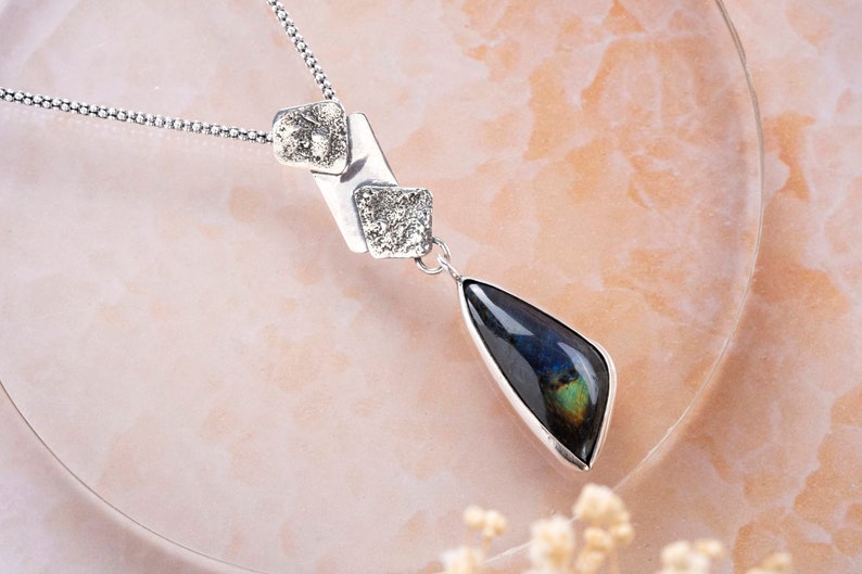 May include: A silver necklace with a teardrop-shaped pendant featuring a dark blue labradorite gemstone. The pendant is attached to a silver chain with two smaller silver squares.