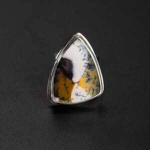 Puede incluir: Anillo de plata con una piedra natural triangular. La piedra presenta un patr&oacute;n &uacute;nico de tonos blancos, amarillos y marrones. El engaste del anillo es de plata pulida, y la piedra es el punto focal de la joya.