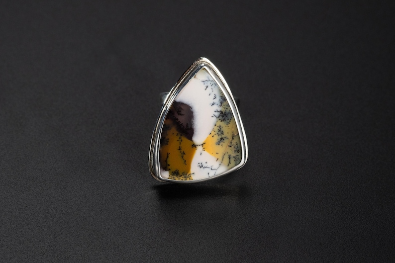 Puede incluir: Anillo de plata con una piedra natural triangular. La piedra presenta un patr&oacute;n &uacute;nico de tonos blancos, amarillos y marrones. El engaste del anillo es de plata pulida, y la piedra es el punto focal de la joya.
