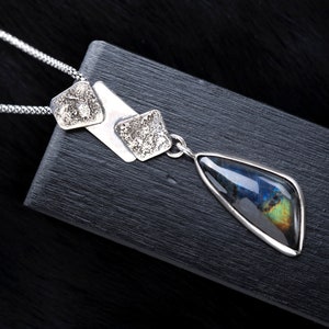 May include: A silver necklace with a teardrop-shaped pendant featuring a iridescent gemstone. The pendant is attached to a chain with three silver square charms.
