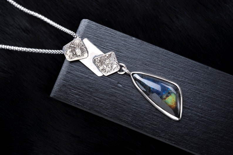 May include: A silver necklace with a teardrop-shaped pendant featuring a iridescent gemstone. The pendant is attached to a chain with three silver square charms.