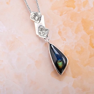 May include: A silver necklace with a teardrop-shaped pendant featuring a dark blue and green iridescent stone. The pendant is attached to a silver chain with three small silver charms.