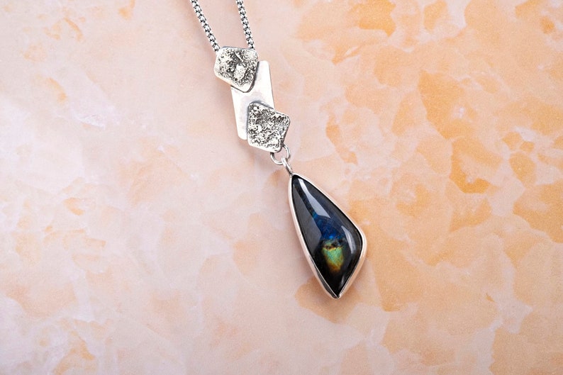 May include: A silver necklace with a teardrop-shaped pendant featuring a dark blue and green iridescent stone. The pendant is attached to a silver chain with three small silver charms.