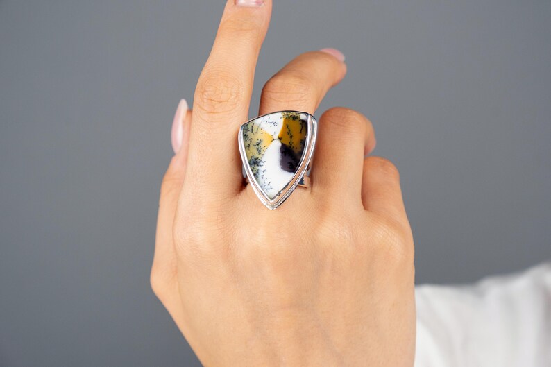 Puede incluir: Un anillo de plata con una piedra grande en forma de escudo. La piedra presenta un patr&oacute;n &uacute;nico de blanco, amarillo y negro. El anillo se muestra en una mano, destacando el dise&ntilde;o y los colores de la joya sobre un fondo gris.