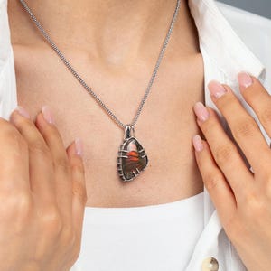 May include: A silver pendant necklace with a unique, irregularly shaped pendant. The pendant features a colorful, layered stone with shades of orange, brown, and red. The necklace is worn over a white shirt.