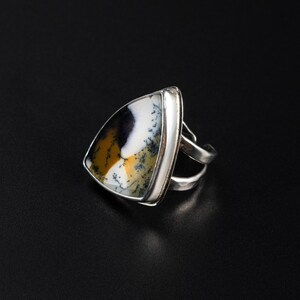 Puede incluir: Un anillo de plata con una piedra triangular que presenta un patr&oacute;n &uacute;nico de tonos negros, blancos y amarillo-marr&oacute;n. El anillo tiene un dise&ntilde;o de doble banda y est&aacute; colocado sobre un fondo oscuro, resaltando los colores de la piedra.