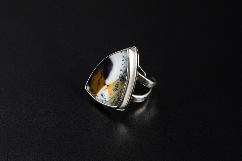 Puede incluir: Un anillo de plata con una piedra triangular que presenta un patr&oacute;n &uacute;nico de tonos negros, blancos y amarillo-marr&oacute;n. El anillo tiene un dise&ntilde;o de doble banda y est&aacute; colocado sobre un fondo oscuro, resaltando los colores de la piedra.