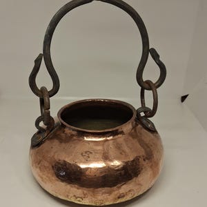 May include: A hammered copper pot with a rounded body and a wide opening. It features a dark, weathered iron handle assembly attached to the sides with metal loops, topped by a circular iron ring for hanging or carrying the vessel securely.