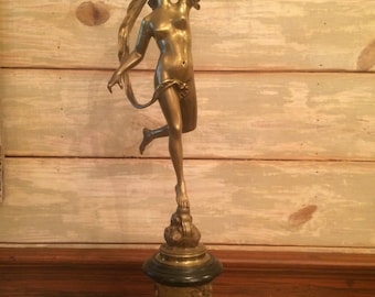 Op de afbeelding: Bronzen beeld van een naakte vrouw die een voorwerp omhoog houdt. Het beeld staat op een zwarte en gouden voet met gedetailleerde reliëfsnijwerk. Het beeld heeft vloeiend haar en bevindt zich in een dynamische pose.
