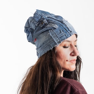 May include: A blue denim beanie hat made from repurposed jeans. The hat features visible stitching, patches, and a small red embroidered detail. The hat is worn by a person with long brown hair.