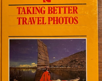 Pode incluir: Um livro amarelo com o título "Taking Better Travel Photos" e o texto "The Kodak Library of Creative Photography". A capa apresenta uma fotografia de uma pessoa com um poncho vermelho em um barco em um lago.