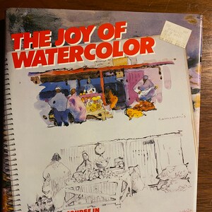 Op de afbeelding: Een boek getiteld "The Joy of Watercolor" met een aquarel-illustratie van een marktscène op de omslag. Het boek is een complete cursus in aquarel schilderen waarbij je je schetsboek als werkboek gebruikt, door David Millard.