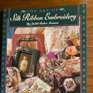 Könnte beinhalten: Das Buchcover "Die Kunst der Seidenband-Stickerei" zeigt eine Vielzahl von farbenfrohen Seidenband-Stickereiprojekten, darunter ein gerahmtes Bild, eine kleine Geldbörse und ein Dekokissen.