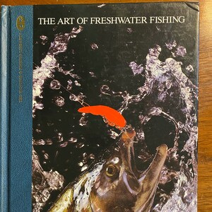 Puede incluir: Un libro titulado "The Art of Freshwater Fishing" con una imagen de primer plano de un pez saltando fuera del agua con un señuelo rojo en la boca.