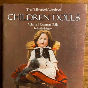 May include: Book cover for "Children Dolls: Volume I German Dolls" by Mildred Seeley. The cover features a color illustration of a doll wearing a red and white dress, sitting on a wooden chair. The doll has blonde hair and blue eyes. The book title is in large, bold letters.