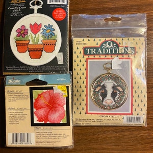 Op de afbeelding: Drie geteld kruissteekpakketten met een koe gezicht, een hibiscusbloem en bloempotten. Elke set bevat een frame, stof, garen, naald, instructies en een diagram. Gemaakt in China.