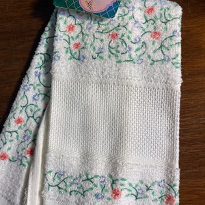 May include: White kitchen towel with a floral embroidery design in shades of pink, green, and blue. The towel has a fringed edge and a tag that reads "Clara Craft".