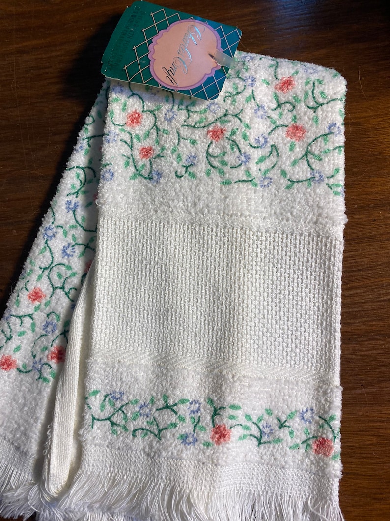 May include: White kitchen towel with a floral embroidery design in shades of pink, green, and blue. The towel has a fringed edge and a tag that reads "Clara Craft".