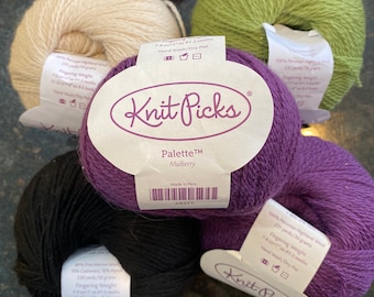 Puede incluir: Cinco ovillos de lana en diferentes colores, incluyendo negro, blanco, verde, morado y un morado oscuro. La lana está etiquetada como "Knit Picks Palette™ Mulberry" y "Made in Peru".
