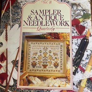 Könnte beinhalten: Ein Magazin mit dem Titel Sampler & Antique Needlework Quarterly Vol. 8, das einen gerahmten Kreuzstich-Sampler mit Alphabet, Zahlen, Vögeln und Blumenmotiven von 1992 zeigt, umgeben von verschiedenen Nähutensilien, Spitze und Stoffresten.