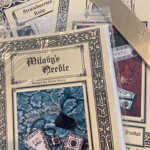 May include: Close-up of cross-stitch patterns and supplies. The main pattern is titled "Milady's Needle" with a floral design and the text "Designs By Gloria Moore." Other patterns include "Strawberries Rule" and "Pocket." A metal ruler is also visible.