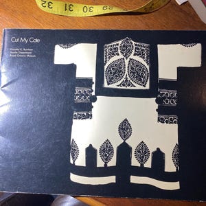May include: A black and white book cover with the title "Cut My Cote" and the text "Dorothy K. Burnham, Textile Department, Royal Ontario Museum". The cover features a black and white silhouette of a traditional garment with intricate patterns.