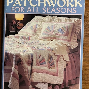 Op de afbeelding: Een knutselboekje met de titel "Patchwork voor alle seizoenen" met een foto van een quilt op een bed. Het boekje is van Leisure Arts en heeft nummer 1585. De prijs is $ 9,95. De quilt is gemaakt van roze, witte en blauwe stof en heeft een gerimpelde rand. De tekst "Vijftien projecten" staat onderaan het boekje.