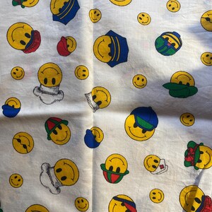 May include: White fabric with a repeating pattern of yellow smiley faces wearing various hats and accessories. The smiley faces are in different sizes and orientations, with blue, red, and green accents.
