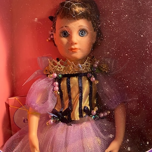 Może przedstawiać: Lalka porcelanowa w fioletowej i złotej sukience z koroną. Lalka ma niebieskie oczy i brązowe włosy.