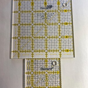 May include: Two clear acrylic Omnigrid rulers with yellow grid lines and measurements. The larger ruler is square, and the smaller one is rectangular. Both have the Omnigrid logo and text indicating U.S. patent information. The rulers are designed for quilting and crafting.
