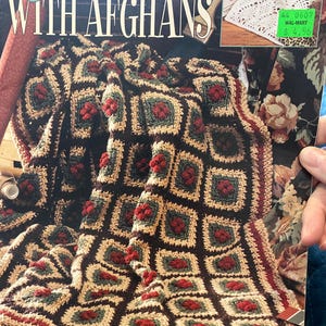 May include: A book cover titled "Decorate with Afghans" featuring a close-up of a crocheted afghan. The afghan has a square pattern with red, green, and tan squares on a dark brown background. The book cover also shows a white lace doily.
