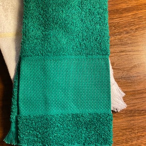 Puede incluir: Una toalla verde doblada con un tejido texturizado y detalles de flecos. La toalla tiene una secci&oacute;n central tejida y una textura esponjosa y absorbente en la parte superior e inferior. Una toalla blanca es parcialmente visible a la izquierda.