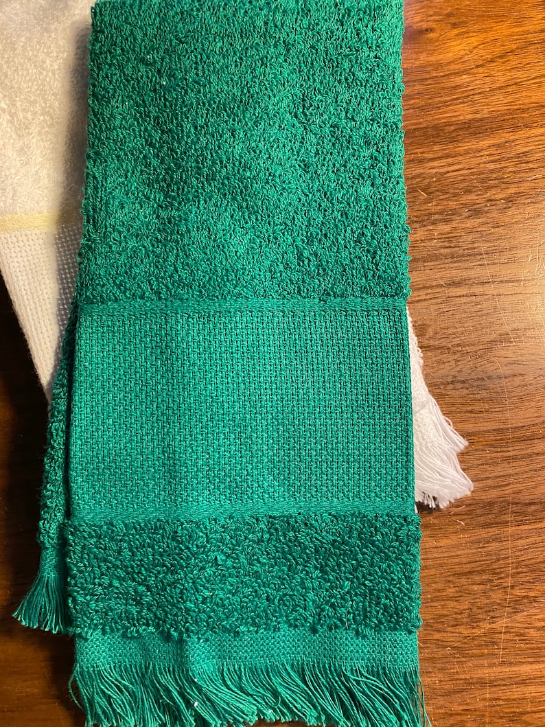 Puede incluir: Una toalla verde doblada con un tejido texturizado y detalles de flecos. La toalla tiene una secci&oacute;n central tejida y una textura esponjosa y absorbente en la parte superior e inferior. Una toalla blanca es parcialmente visible a la izquierda.