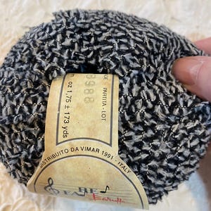 Puede incluir: Una madeja de hilo negro y blanco con una superficie texturizada. Una etiqueta beige adjunta muestra el texto "3988" y "1.75 oz ± 173 yds". La etiqueta también incluye el texto "DISTRIBUITO DA VIMAR 1991 - ITALY".