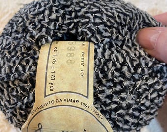 May include: A ball of black and white yarn with a textured surface. A tan label is attached, displaying the text "3988" and "1.75 oz ± 173 yds". The label also includes the text "DISTRIBUITO DA VIMAR 1991 - ITALY".
