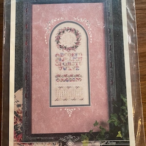 Könnte beinhalten: Ein Kreuzstichmuster mit dem Titel "Memories of Spring" von Emie Bishop. Das Design zeigt einen Blumenkranz, das Alphabet und dekorative Elemente in einer gerahmten Präsentation. Die Farbpalette umfasst Rosa, Grün und verschiedene andere Farben.