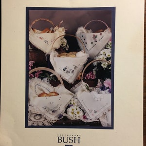 Puede incluir: Una colección de cuatro patrones de punto de cruz con cestas de pan y flores. Los patrones se titulan "Shepherd's Bush" y son del Folio Dos de Printworks.
