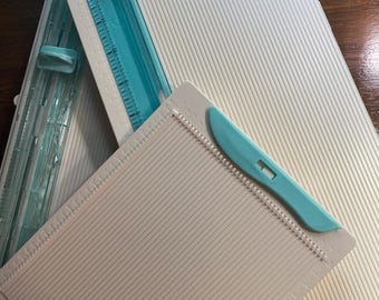 Puede incluir: Un conjunto de herramientas de manualidades en papel blancas y turquesas, que incluyen una tabla de puntuación con una cuchilla de puntuación turquesa y una cizalla de papel con una guía de corte turquesa. Para cortar y marcar papel con precisión.