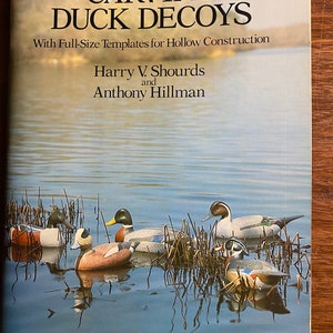 Puede incluir: Portada de libro con el título "Carving Duck Decoys" con plantillas de tamaño real para la construcción hueca. Los autores son Harry V. Shourds y Anthony Hillman. La portada presenta una pintura de cinco señuelos de pato tallados en un entorno de lago.