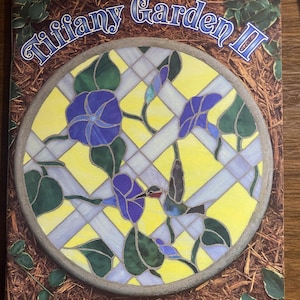 May include: A book titled Tiffany Garden II featuring a circular stained glass mosaic design with purple morning glory flowers, green leaves, and a hummingbird against a yellow lattice background, with the subtitle Book II: Mosaic Tables, Stepping Stones and Bricks by Jean Bishop and Julie Bishop-Day.