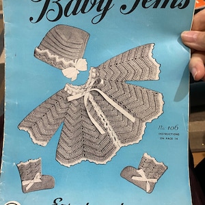 以下が含まれることがあります： ベビー用の手編みパターンブックレットのヴィンテージ。表紙には、グレーと白のクロシェで編まれたベビー服が描かれており、白いリボンが付いています。タイトルは「Baby Gems」で、テキストには「Easy to make Crochet and Knitted Baby Garments by Nell Armstrong」と書いてあります。