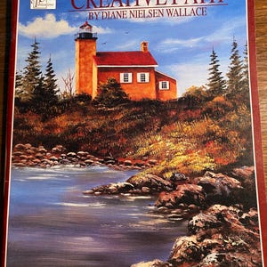May include: A book titled "Creative Path" by Diane Nielsen Wallace, featuring a painting of a red brick lighthouse on a rocky shore with a blue body of water in the foreground.