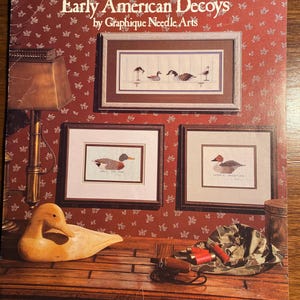 Puede incluir: Un folleto vintage de patrones de punto de cruz titulado "Early American Decoys" de Graphique Needle Arts. La portada presenta un señuelo de pato de madera, una mesa de madera con una lámpara y dos patrones de punto de cruz enmarcados de patos. El folleto está etiquetado como "Leaflet #12".