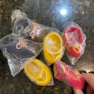 Puede incluir: Variedad de zapatos de muñeca en miniatura, envueltos en plástico transparente. Los zapatos son de varios colores: negro, amarillo y rojo. Algunos tienen cordones, otros detalles en forma de corazón. Sobre una superficie oscura y moteada.