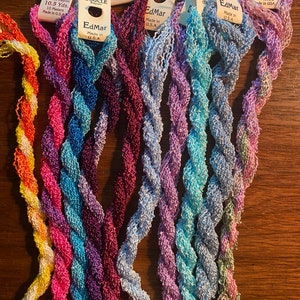 以下が含まれることがあります： 黄色、ピンク、青、紫、水色など、さまざまな色のブークレ糸の糸巻き8個の詰め合わせ。各糸巻きには、「EdMar」というブランド名と、「Boucle」という糸の種類、色番号、糸の長さが表示されています。