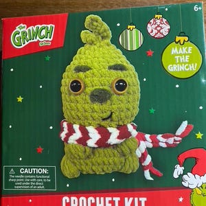May include: A Dr. Seuss The Grinch crochet kit box featuring a green crocheted character with large brown eyes and a red and white striped scarf. The packaging includes text stating Make The Grinch, Crochet Kit, and lists contents like 43.2 yards of yarn.