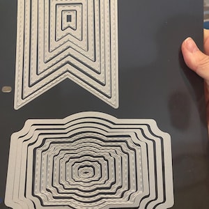 Puede incluir: Un conjunto de troqueles de metal plateado con forma de pancarta y etiqueta. Los troqueles de pancarta tienen un diseño de borde cosido. Los troqueles de etiqueta tienen un borde festoneado. Los troqueles están sobre un fondo negro.