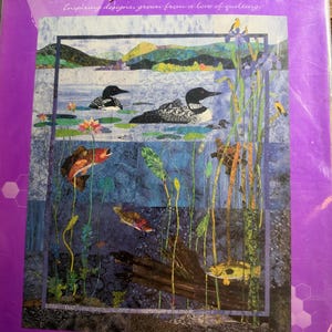 May include: A quilt pattern package titled Day at the Lake featuring a lake scene with loons, fish, turtles, and aquatic plants. The purple packaging includes the text Day at the Lake, Inspiring designs, grown from a love of quilting, and bee creative.